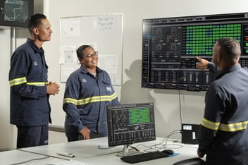 Trabalhadores com uniformes de segurança em uma sala de controle, com grandes telas que exibem dados técnicos e controles do sistema.