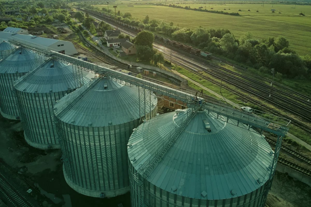 Vista aérea de silos e armazéns agrícolas com telhados metálicos azuis ao lado de trilhos de trem e paisagem rural com campos e árvores ao fundo.